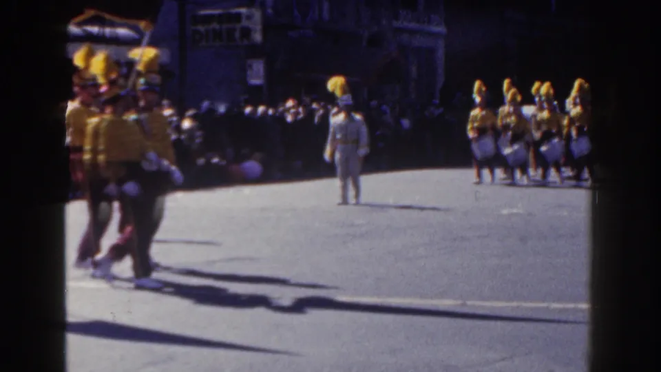 1962 New York City home-movie frame showing a yellow-and-white drumline approaching after the red-uniformed band section.