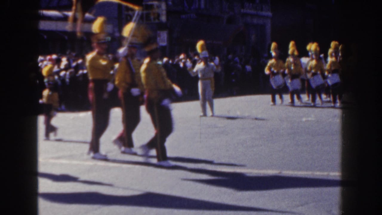 In 1962 New York City footage, uniformed marching band members with yellow plumes move past a sidewalk crowd on a sunlit street.