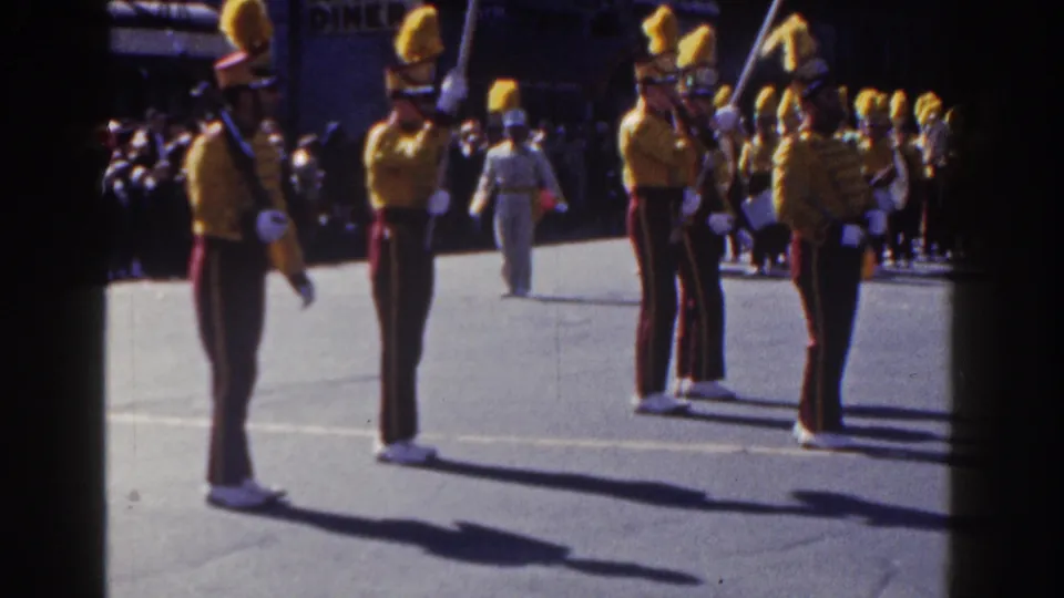 Yellow-uniformed marching band members with tall plumes are seen closer to camera during a 1962 New York City parade, with spectators behind them.
