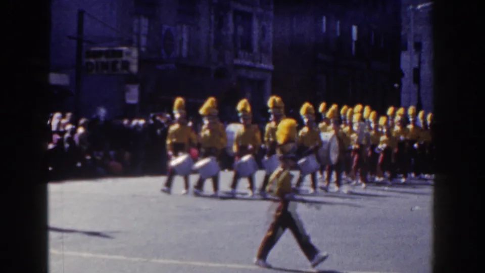 The final frame widens to a drum line and yellow-uniformed marchers crossing a New York City street, with spectators along the shaded sidewalk.