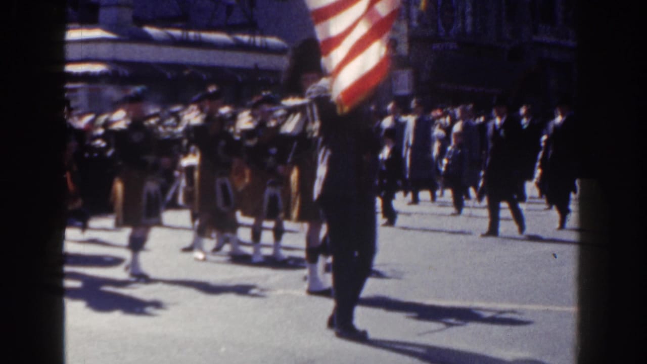 Kilted pipe band marches on a sunny city street as an American flag passes in the foreground.