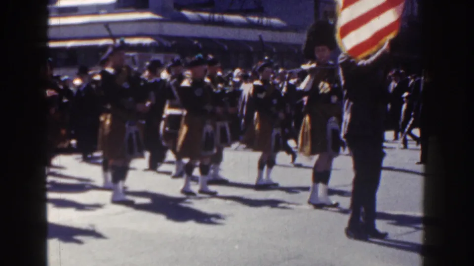 Kilted bagpipers and drummers march in formation on a bright New York City street, with an American flag visible at the right edge.