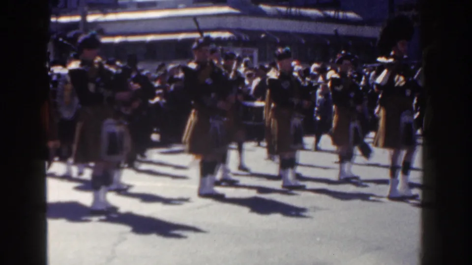 Home-movie frame from the 1962 New York City parade showing kilted band members moving past the camera in strong daylight.