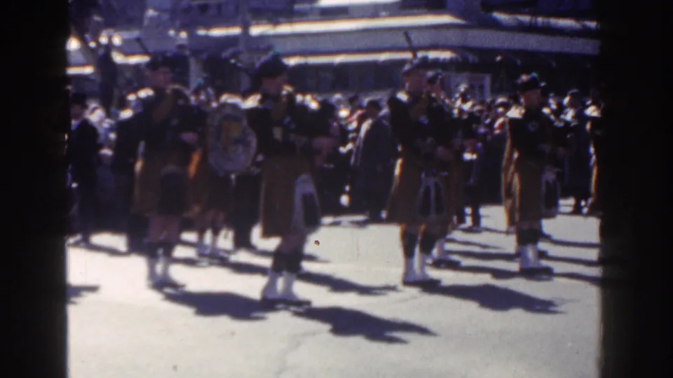 A 1962 New York City parade frame shows kilted drummers and bagpipers passing spectators in front of a large building.