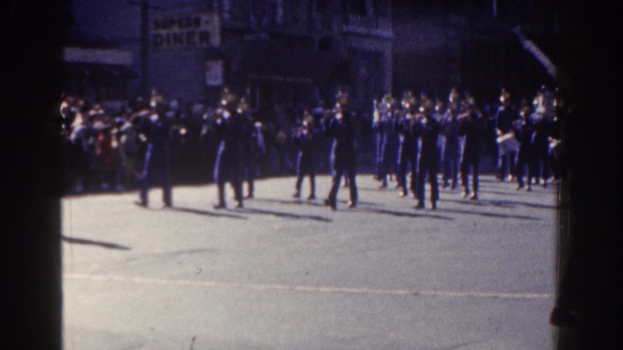 Marching band members cross a city street while spectators watch from the sidewalk.