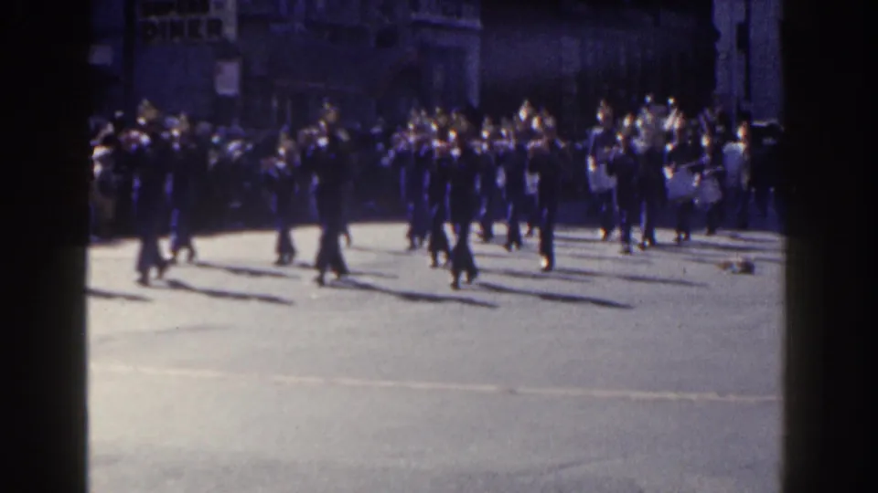 A New York City marching band proceeds through a sunny street scene, with a crowd watching from the sidewalk in the background.