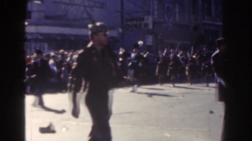 A uniformed armed man appears in the foreground of the 1962 New York City parade scene, with band members and spectators behind him.