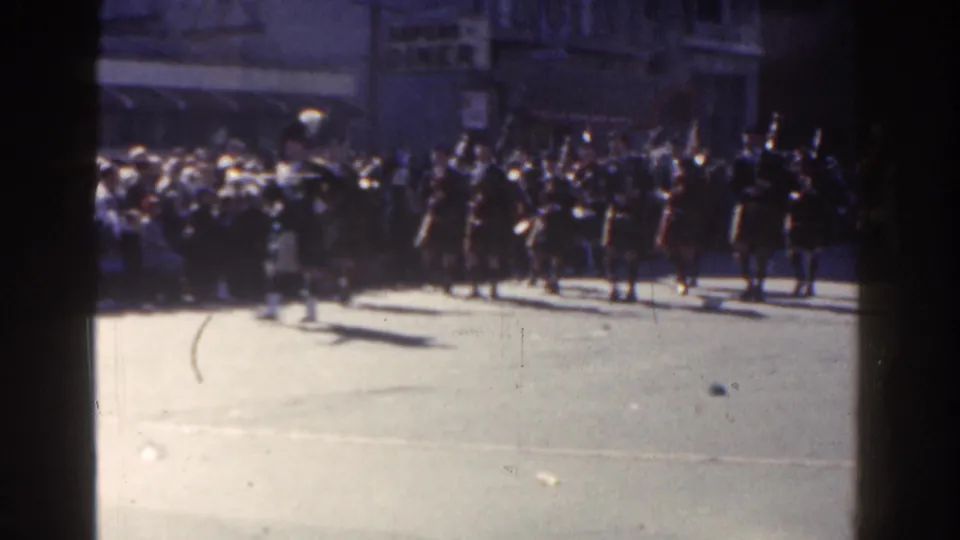 The parade scene continues with band members clustered near the sidewalk and spectators watching across a bright New York City street.