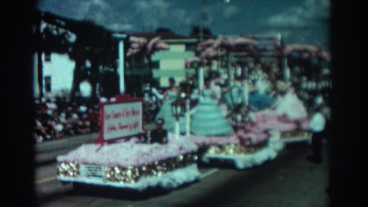 Decorated parade float with pastel-gowned women and a red-bordered sign passing spectators.