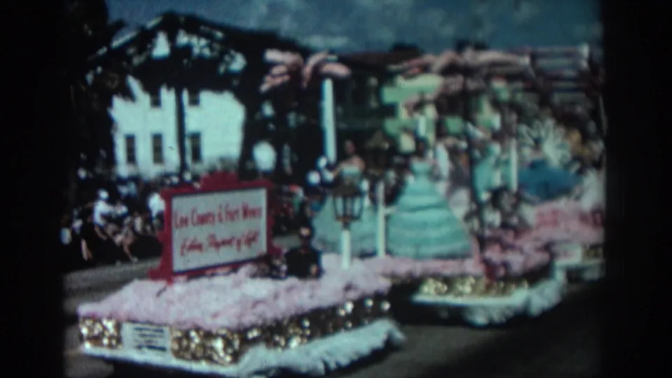 1962 Pasadena frame of a flower-trimmed parade float with young women in full gowns riding past spectators in a home-movie street view.