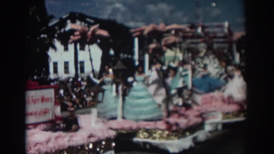 1962 Pasadena home-movie frame with costumed women waving from a pink and gold decorated float during a street parade.