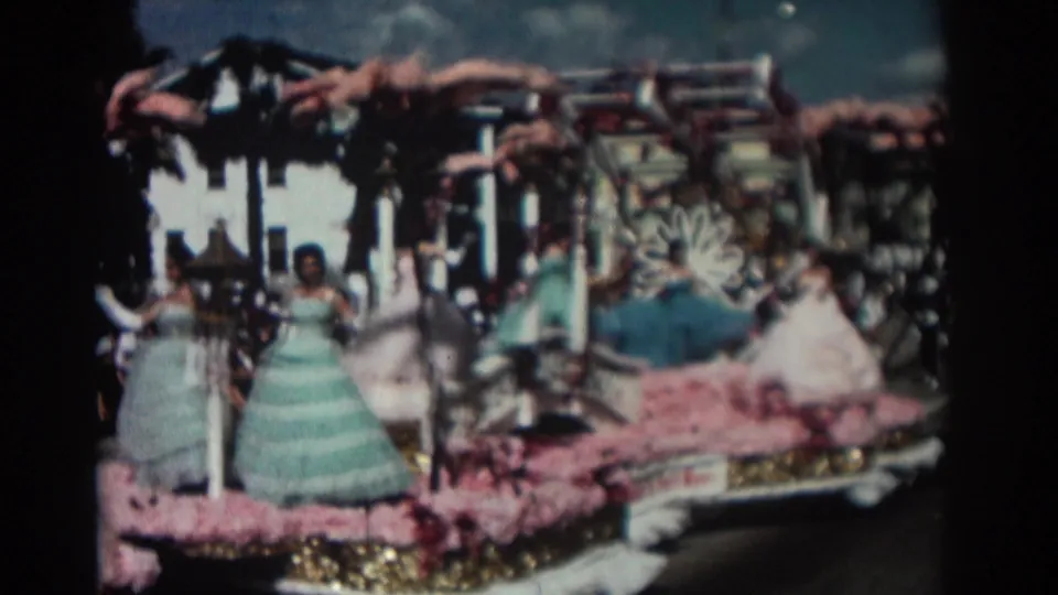 Late shot from the 1962 Pasadena parade clip showing costumed young women on a floral float, with spectators and trees in the background.