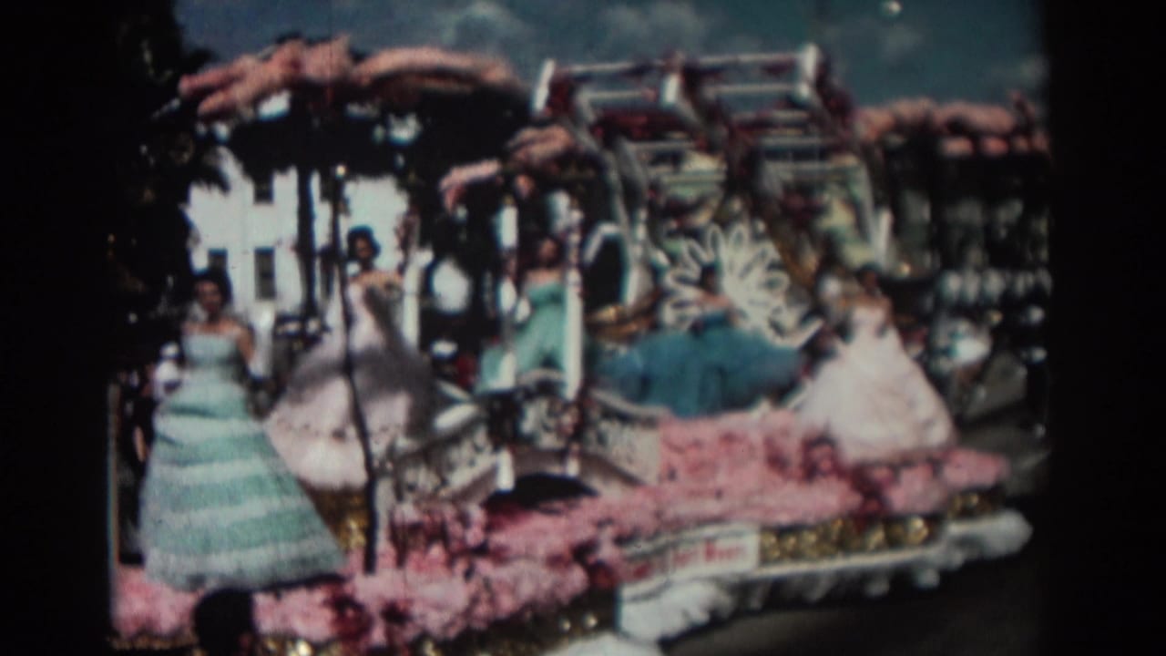 Women in pastel formal gowns ride on a decorated parade float with pink flowers and ornate trim.