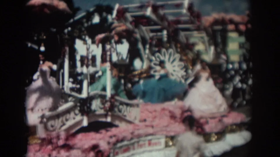 The Pasadena parade float moves through frame, showing costumed women seated or standing among pink flowers, gold trim, and white decorative shapes.