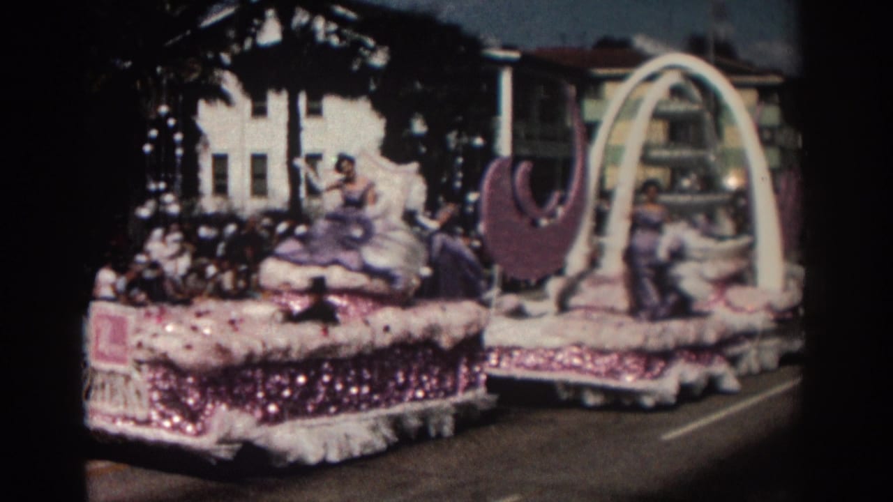 Decorated parade float with women in gowns, pink trim, white arch, and purple crescent forms.