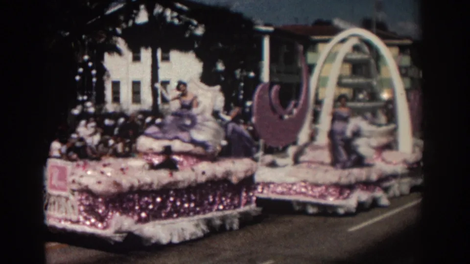 1962 Pasadena parade float with pink sequined trim, white arch, purple crescents, and women in formal gowns on a sunny street.
