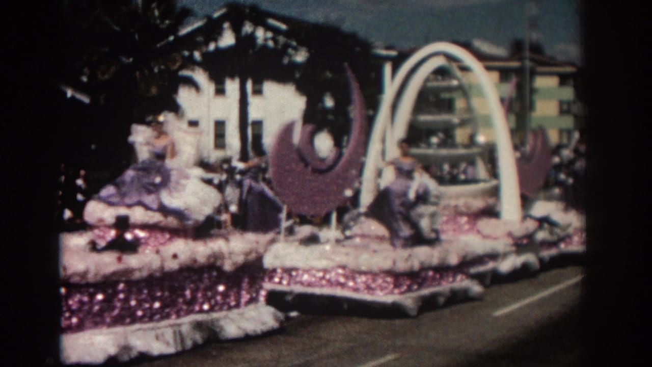 Parade float moving past camera with white arch, purple crescents, and costumed women seated or standing.