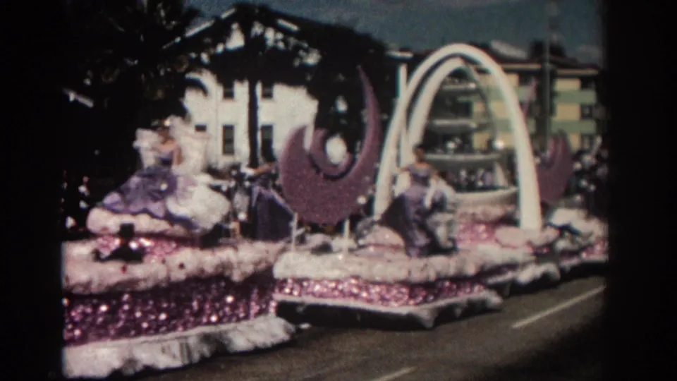 Curbside 8mm view of a Pasadena parade float with a white arch, purple crescent decorations, and women in purple and white gowns.