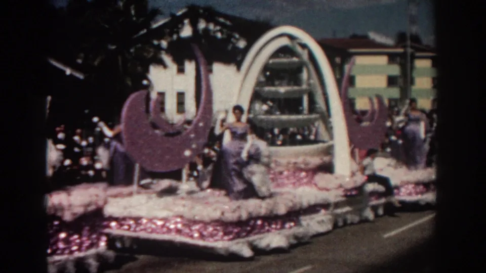1962 Pasadena parade footage showing the side of a decorated float with a large purple crescent, white arch, and women in gowns.