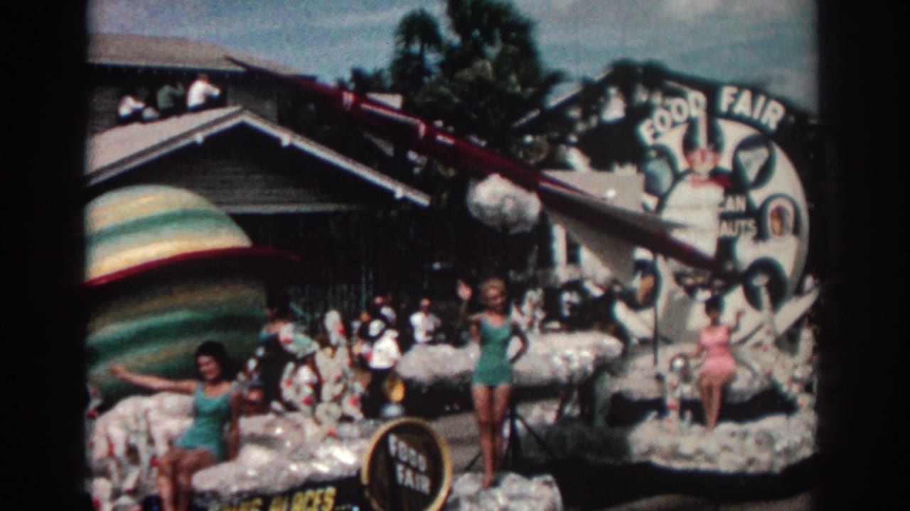 Parade float with rocket and planet decorations, women posing and waving, spectators, and a Food Fair sign.
