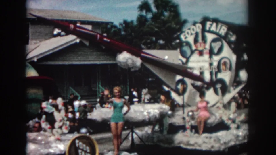 Closer 1962 Pasadena home-movie view of a rocket-themed parade float with women waving near a Food Fair sign.
