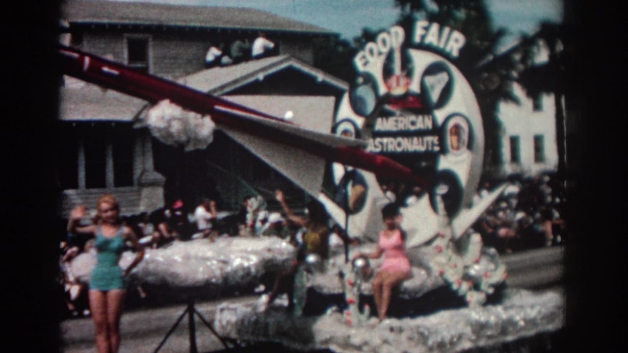 Parade float with astronaut sign, model rocket, women in swimsuits, and spectators along a residential street.