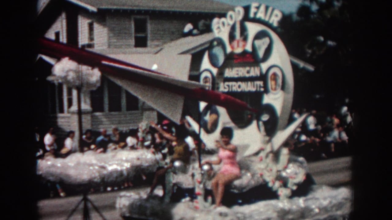Astronaut-themed float with model rocket and seated performer moving past spectators on the street.