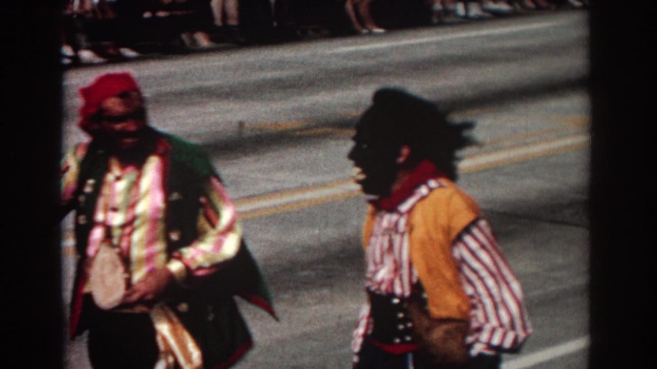 Two costumed parade performers walking on a sunny street with spectators behind them.