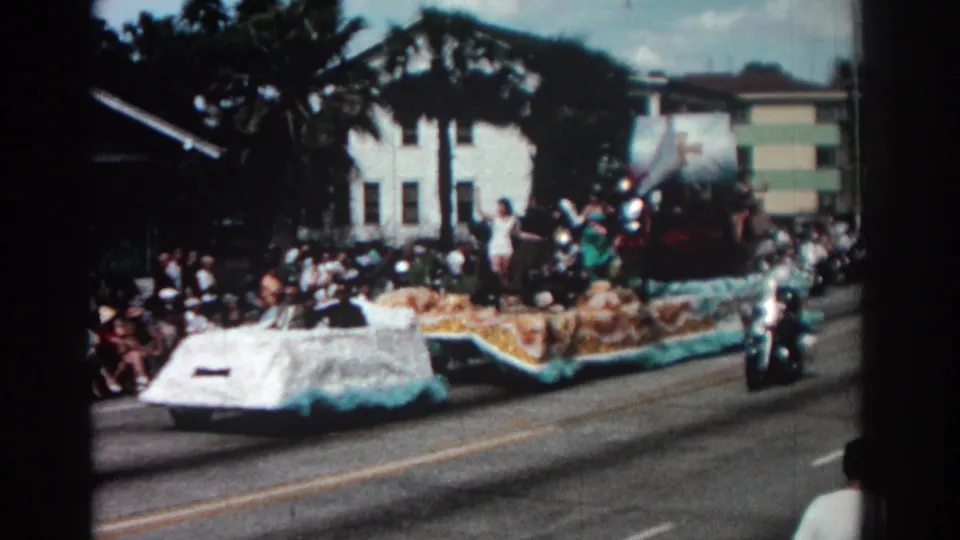 1962 Pasadena parade float with blue trim and yellow decoration passes a curbside crowd beneath palm trees.