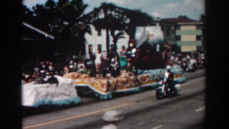 The Pasadena parade float continues down the street with participants aboard and a small vehicle moving alongside the crowd.