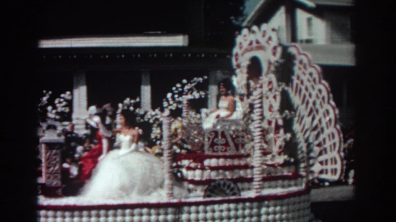 Flower-covered parade float with a woman in a white gown passing spectators on a street.
