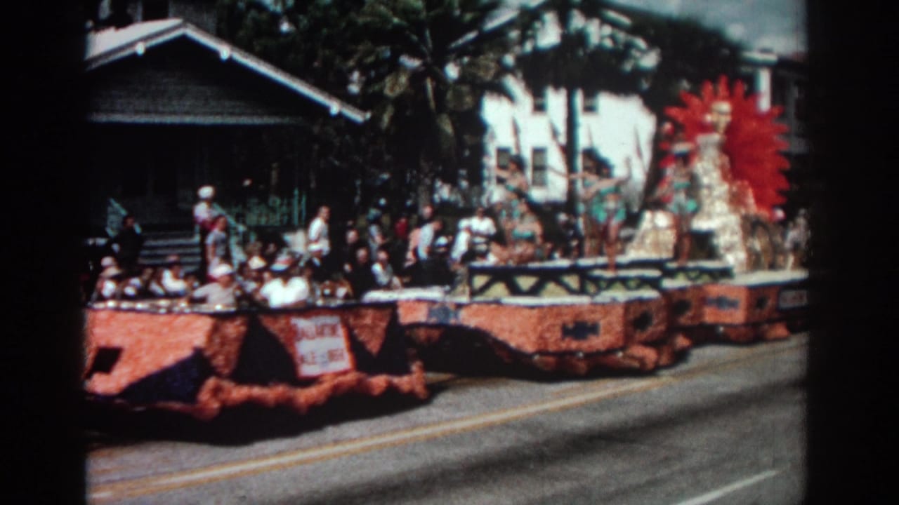 Orange parade float with costumed performers and a red sunburst backdrop moving along a street.
