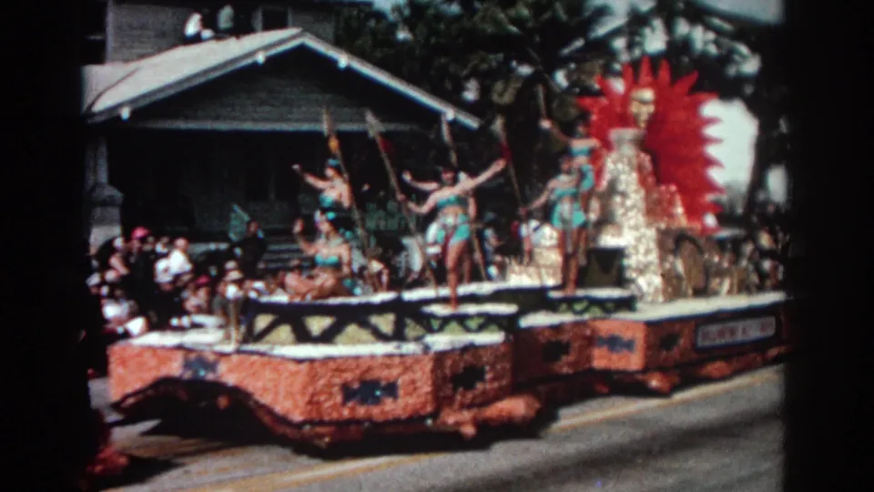 1962 Pasadena parade float showing costumed dancers, a red sunburst decoration, and onlookers beside a residential street.