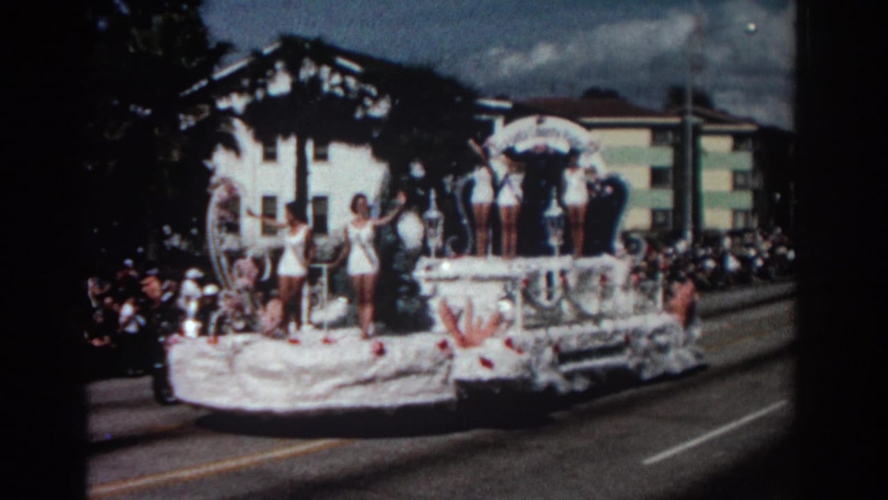 Decorated parade float with women in white swimsuits waving to spectators along a street.