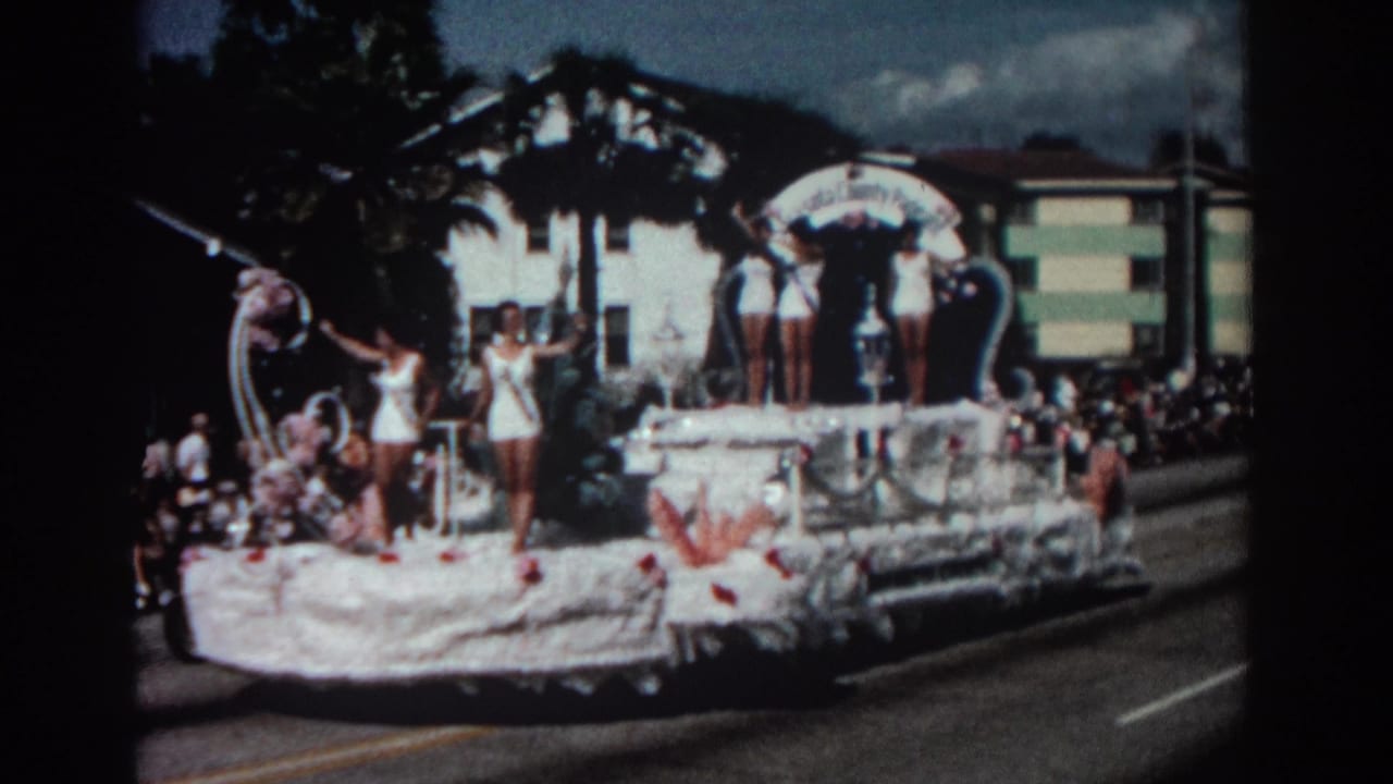 White parade float passing a crowd, with swimsuit-clad women standing and waving.