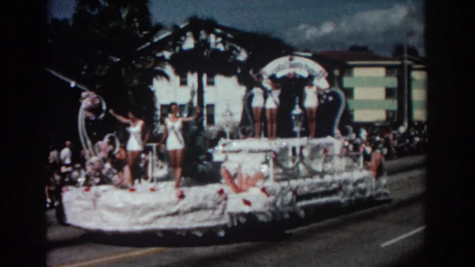 The Pasadena parade float moves along the street with women in white swimsuits and sashes waving from its raised platform.