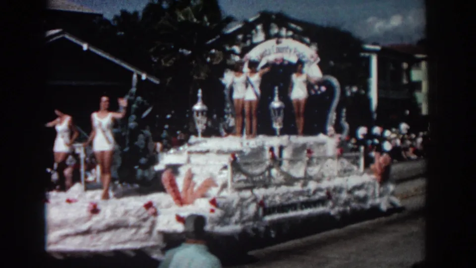Late in the clip, the decorated float continues past the crowd in Pasadena, with women in white swimsuits visible against trees and low-rise buildings.