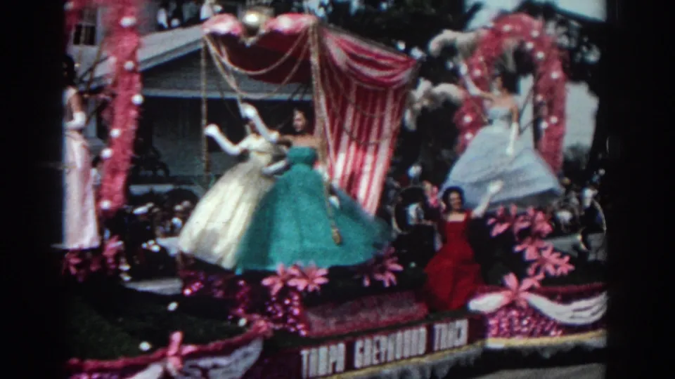 Close home-movie view of a Pasadena parade float with women in formal gowns, pink flowers, and a striped canopy.