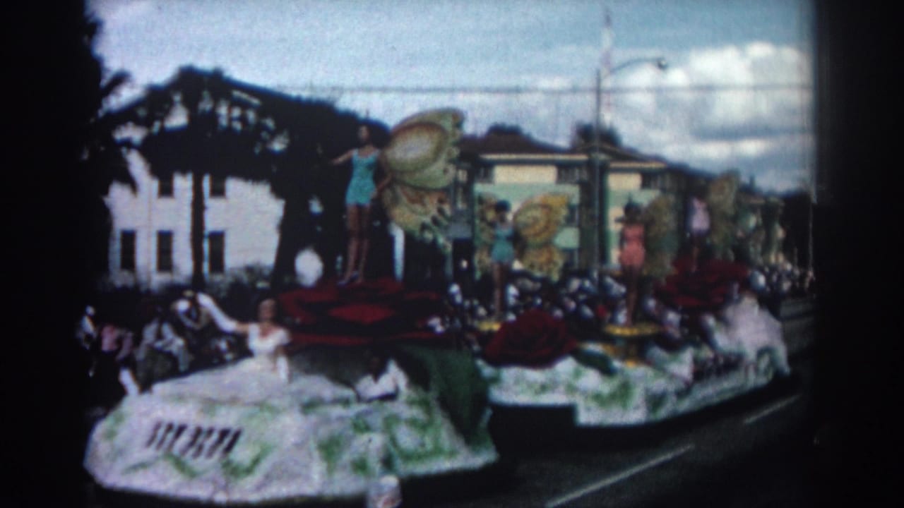 Decorated parade float moves past spectators, with women standing among butterfly wings and red rose forms.