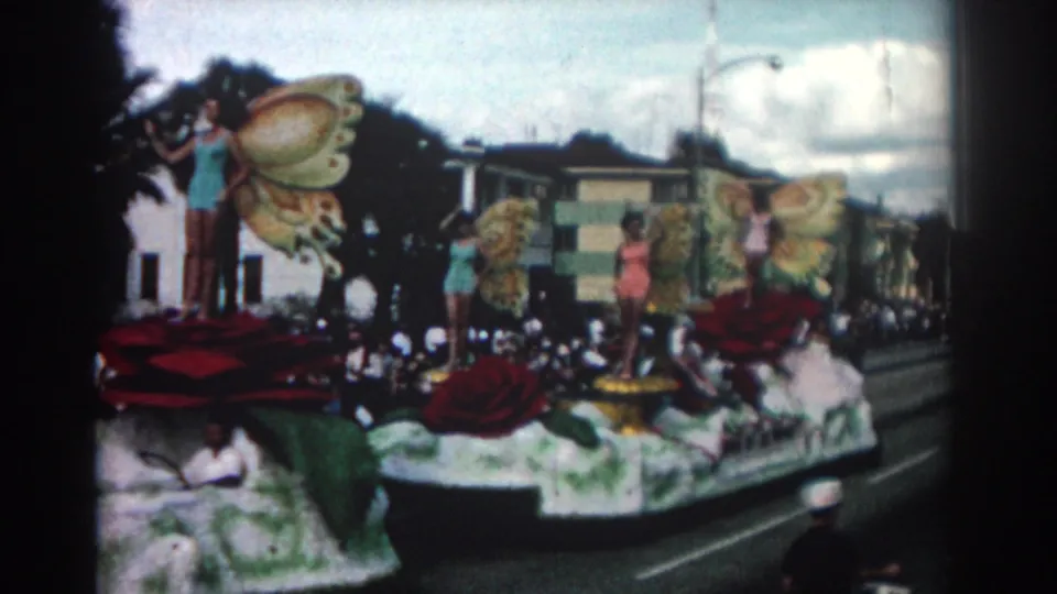 1962 Pasadena parade frame with the float continuing past the camera, showing butterfly-wing props, red roses, women posing, and roadside spectators.