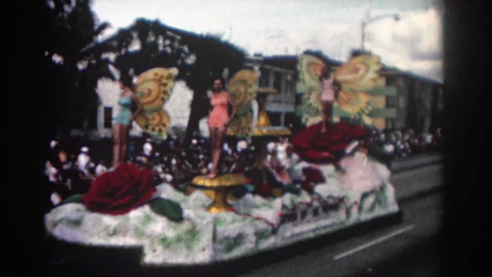 Late shot of the Pasadena parade float with butterfly-costumed women, red rose decorations, crowds, buildings, and cloudy sky.