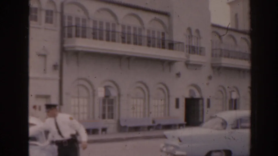 1963 Florida street view with a light facade, arched windows, balcony railings, a uniformed man, and mid-century parked cars.