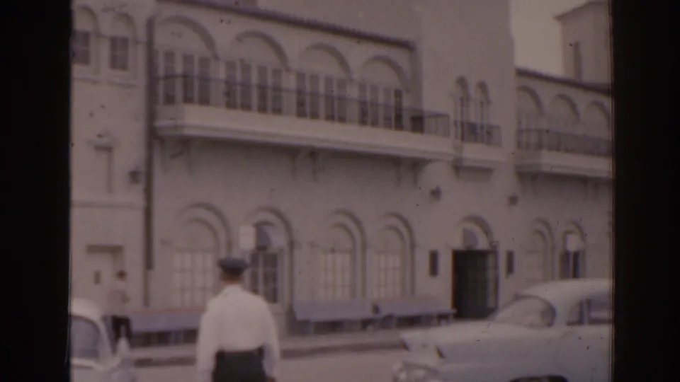 1963 Florida 8mm street frame showing an arched building facade, balcony railings, a uniformed man, and a rounded 1960s car.