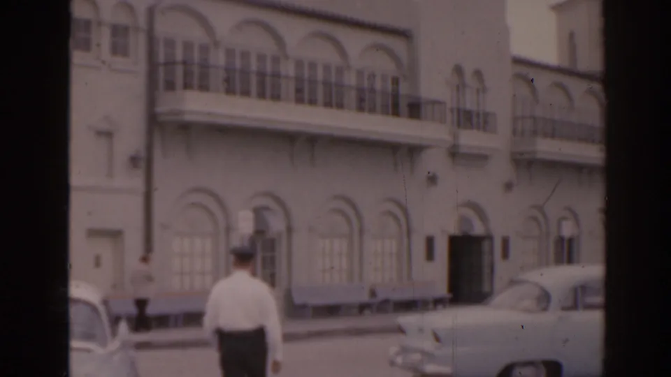 1963 Florida home-movie view of a man walking away beside classic cars and a white arched coastal building.