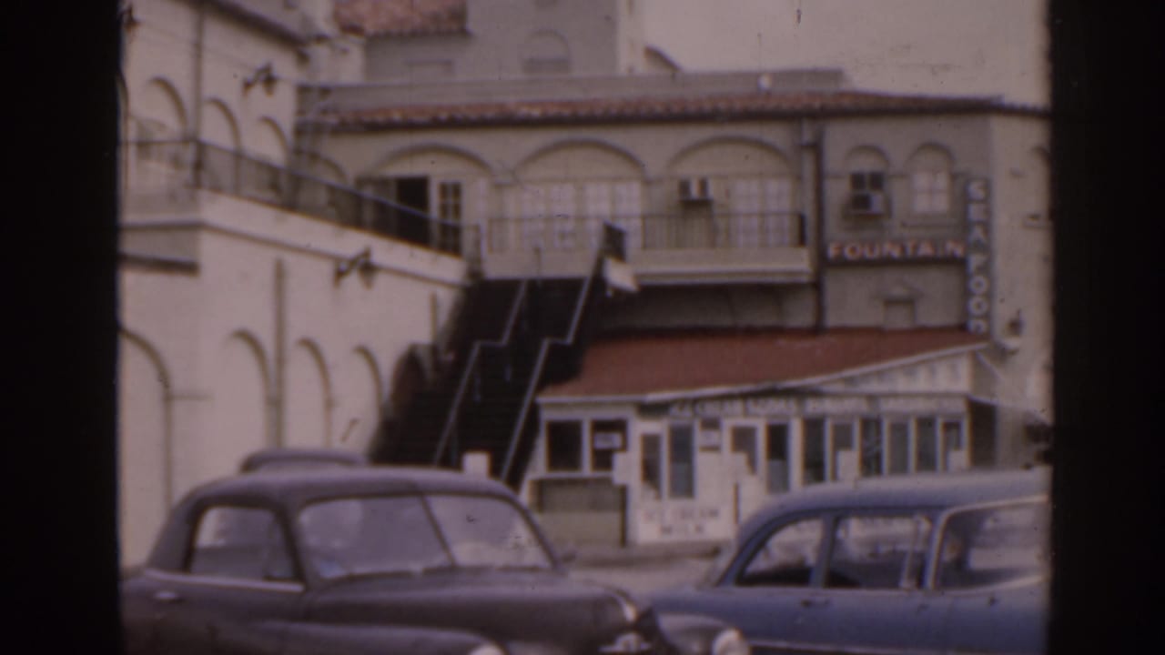 White stucco building with exterior stairs, small storefront, parked cars, and Fountain sign.