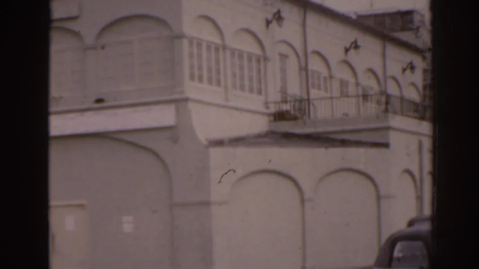 Close 1963 Florida view of a white stucco facade with arched wall details, enclosed windows, balcony rail, and part of a parked car.