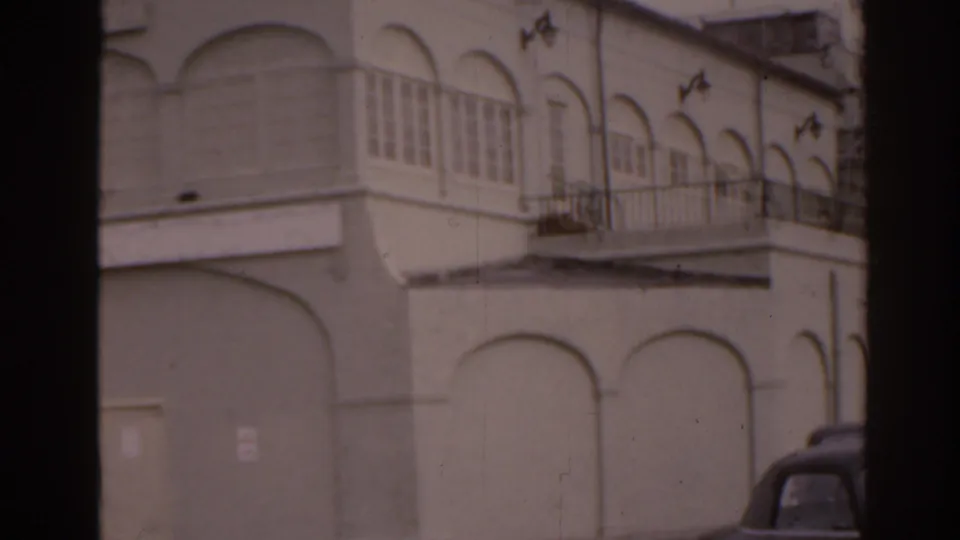 1963 Florida home-movie frame focused on a white stucco building facade, balcony rail, enclosed windows, and part of a parked car.