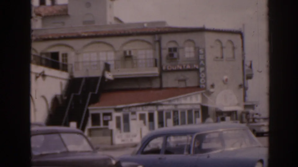 1963 Florida 8mm frame showing an arched two-story building, vertical Fountain sign, small storefront, and parked early 1960s cars.