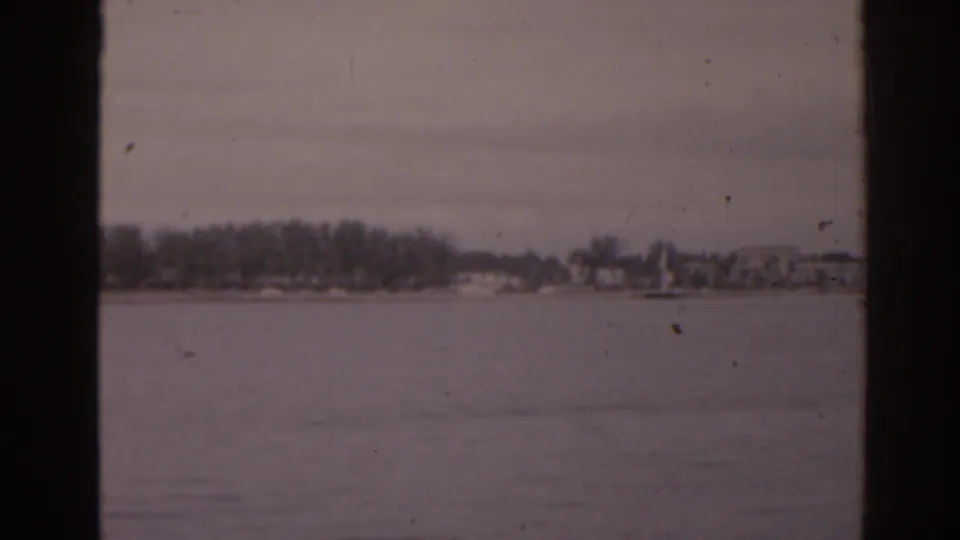 Florida home-movie frame from 1963 showing open water and a distant tree-lined shore with low buildings under a muted sky.