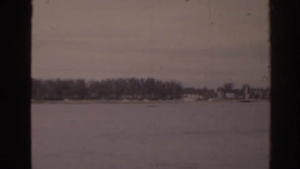 Vintage 8mm frame of a Florida lake with a far tree line and faint buildings under an overcast sky.